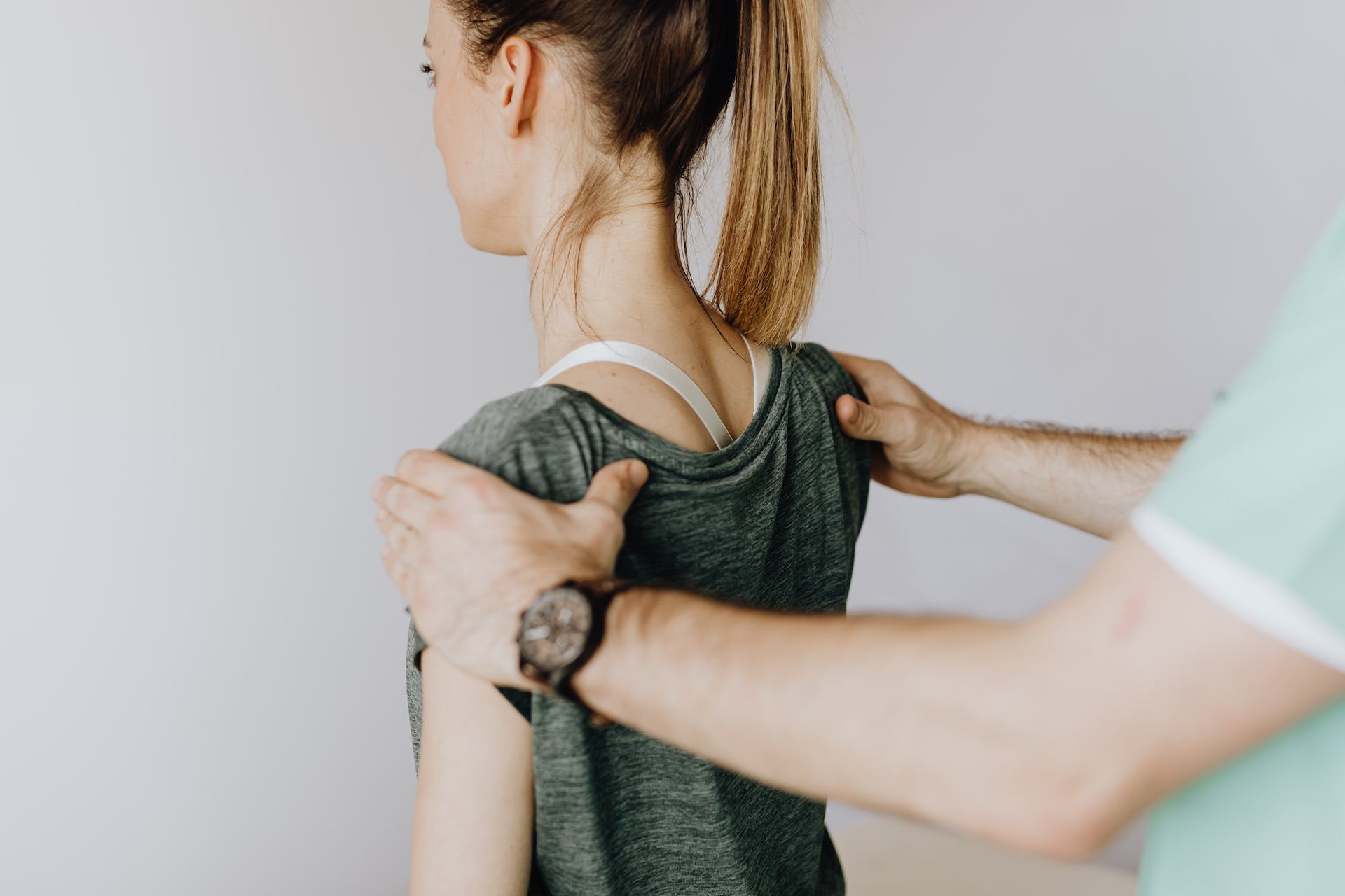 Back view of crop faceless orthopedist in uniform and wristwatch examining shoulder joints of slim female patient in clinic on white background